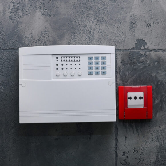 Fire alarm push button and house security system control panel on grey wall, space for text